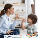 Young woman speech therapist studying together with small kid, learning practice pronunciation exercises with little boy